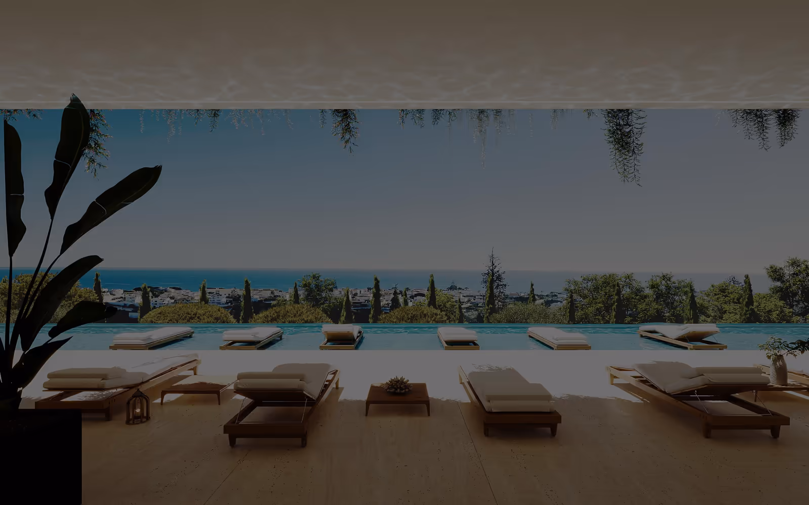 A covered deck with timber loungers facing an infinity pool, the sea and a town beyond.